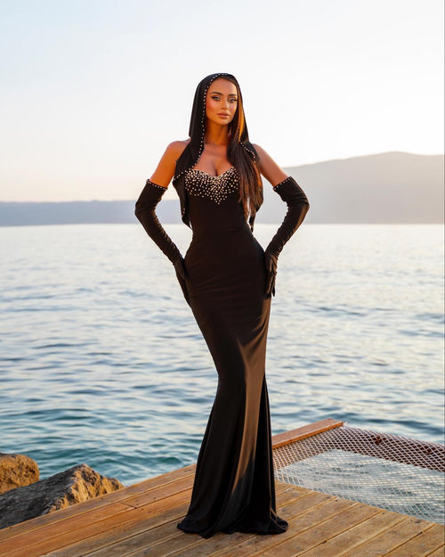 Woman in a black evening gown with gloves standing on a dock by the water.