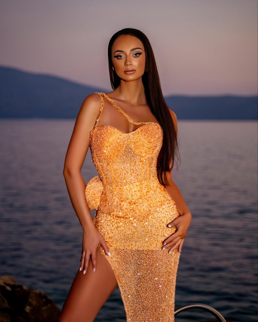 Woman in a gold dress standing by a body of water with mountains in the background