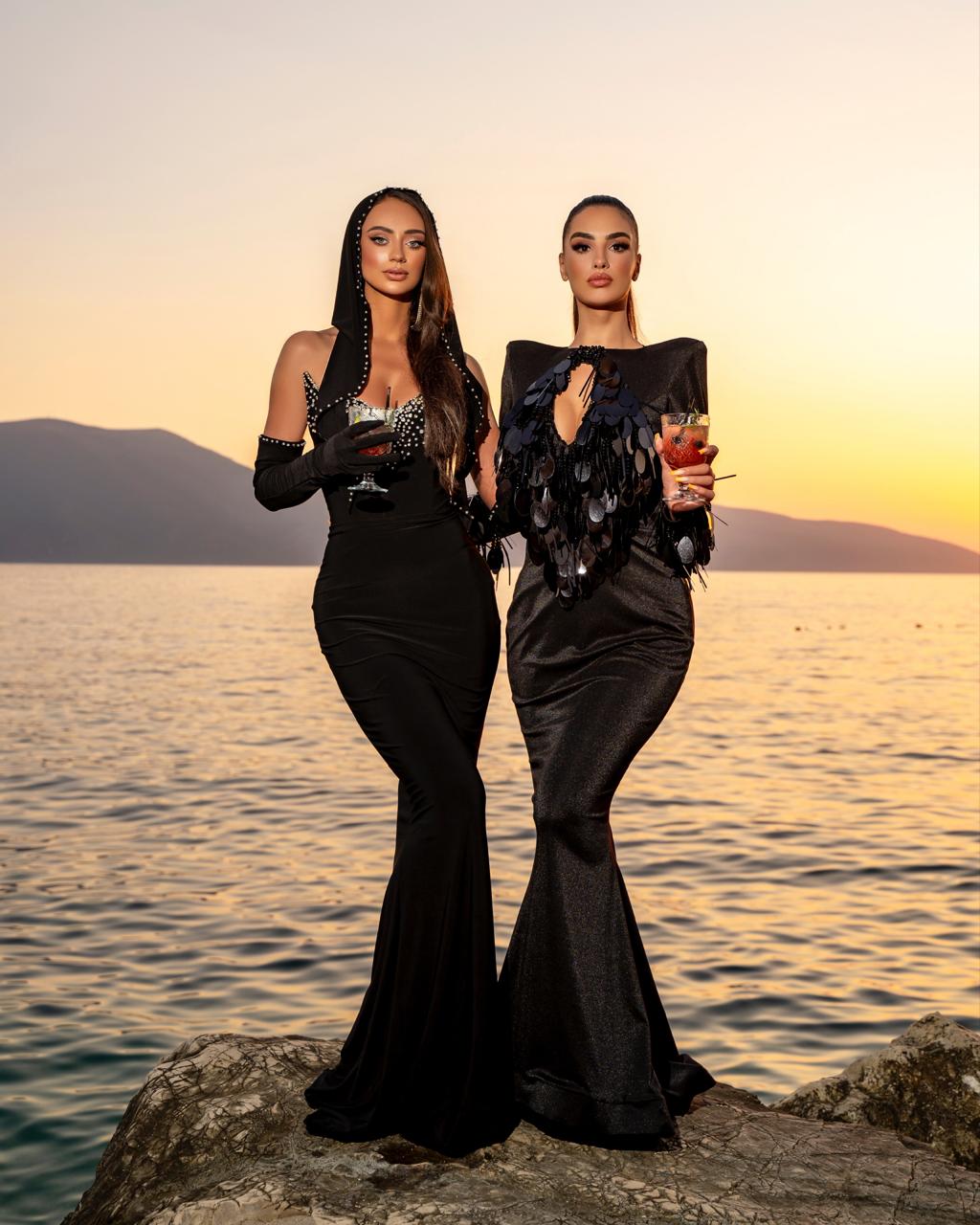 Two women in elegant black evening gowns standing on a rocky shore with a sunset in the background.