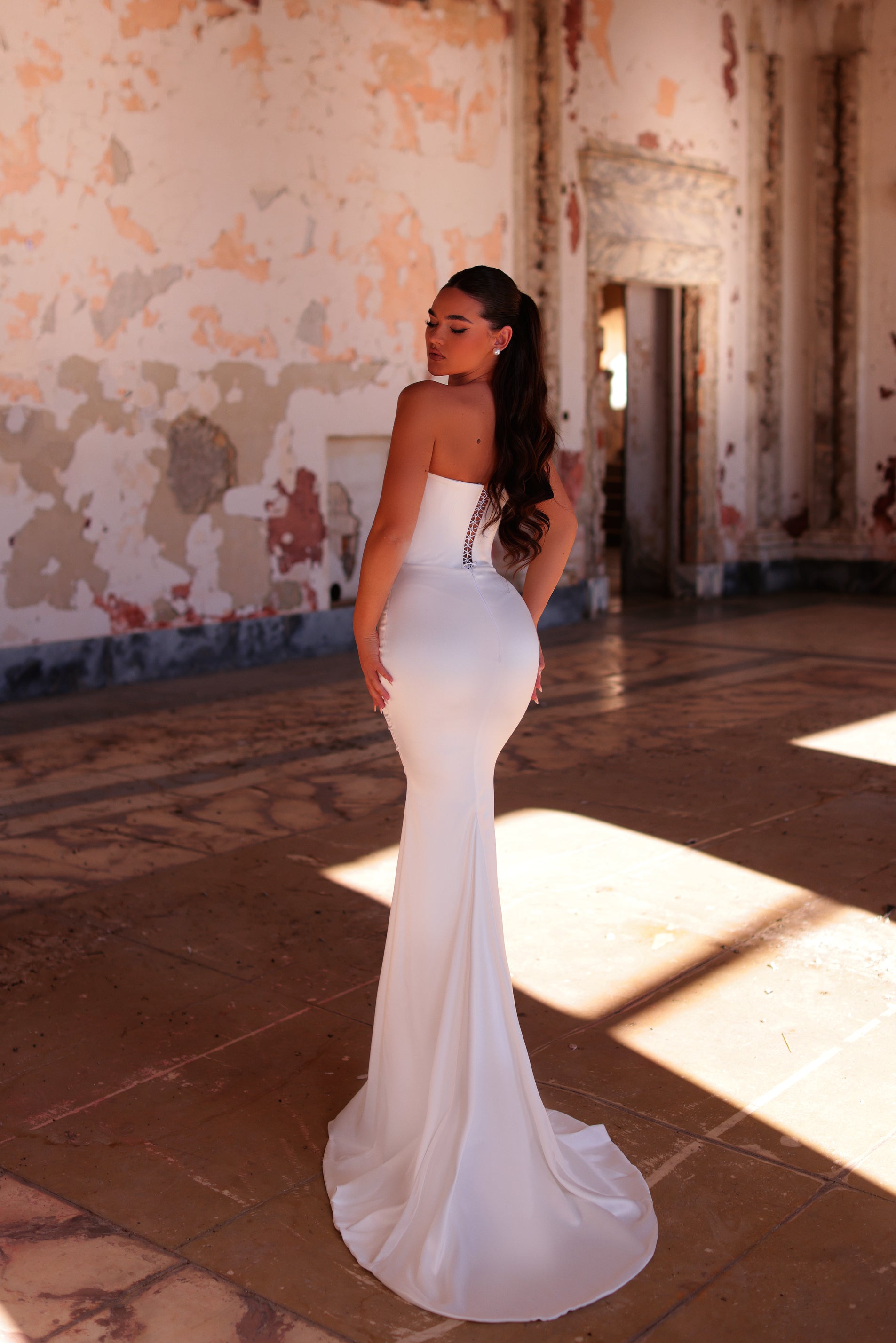 Woman in a white strapless gown standing in an old, sunlit room with peeling walls.