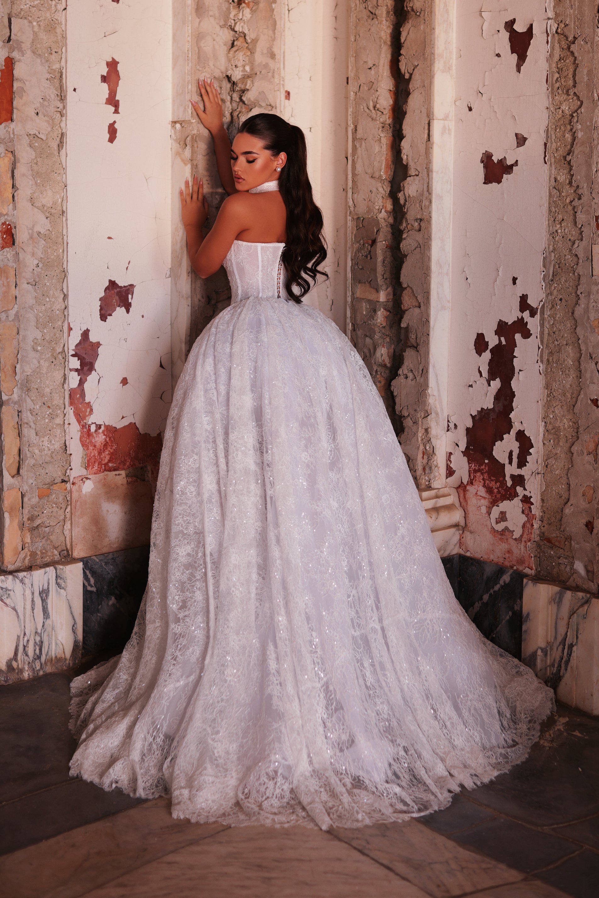 Woman in a white lace wedding dress standing against a textured wall.
