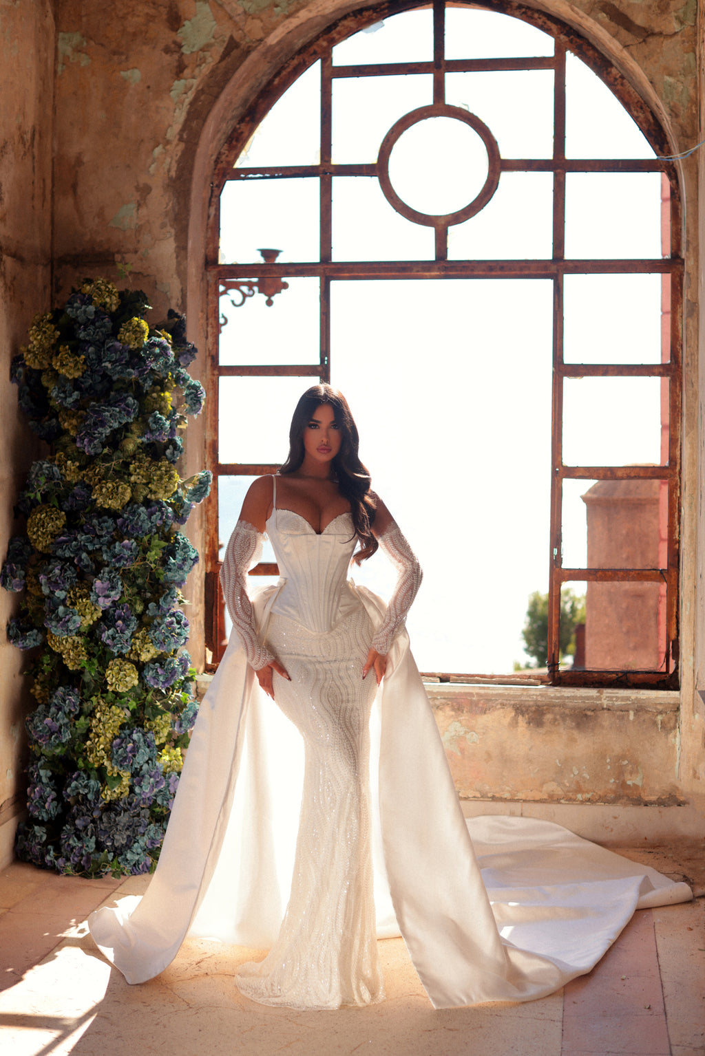 Woman in a white wedding dress standing in a sunlit room with floral decorations.