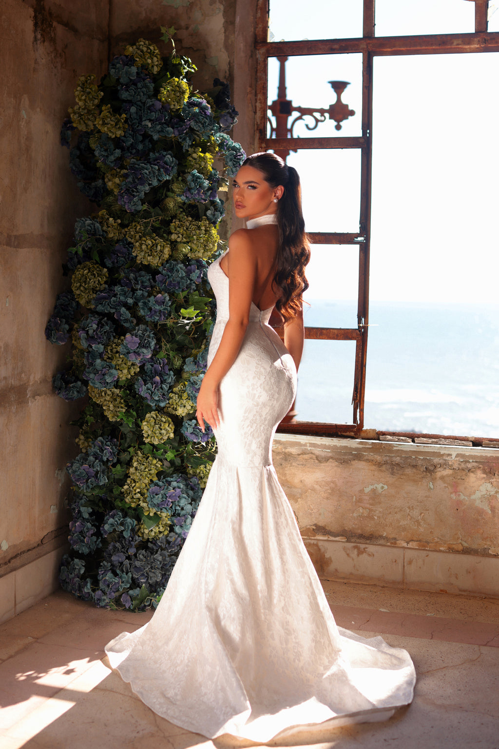 Woman in a white wedding dress standing in a room with floral decorations and large windows.