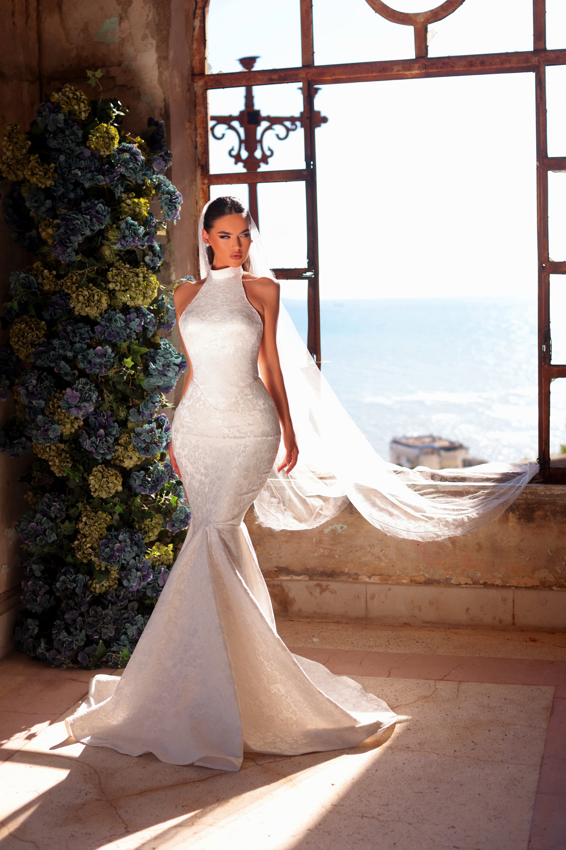 Woman in a white wedding dress standing in a sunlit room with floral decorations.