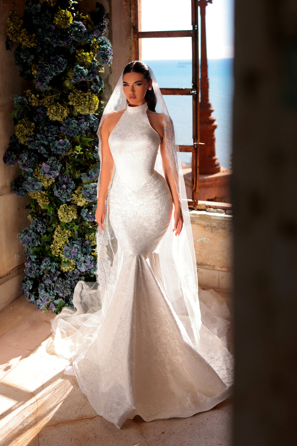 Woman in a white wedding dress standing in a room with floral decorations and a window view.