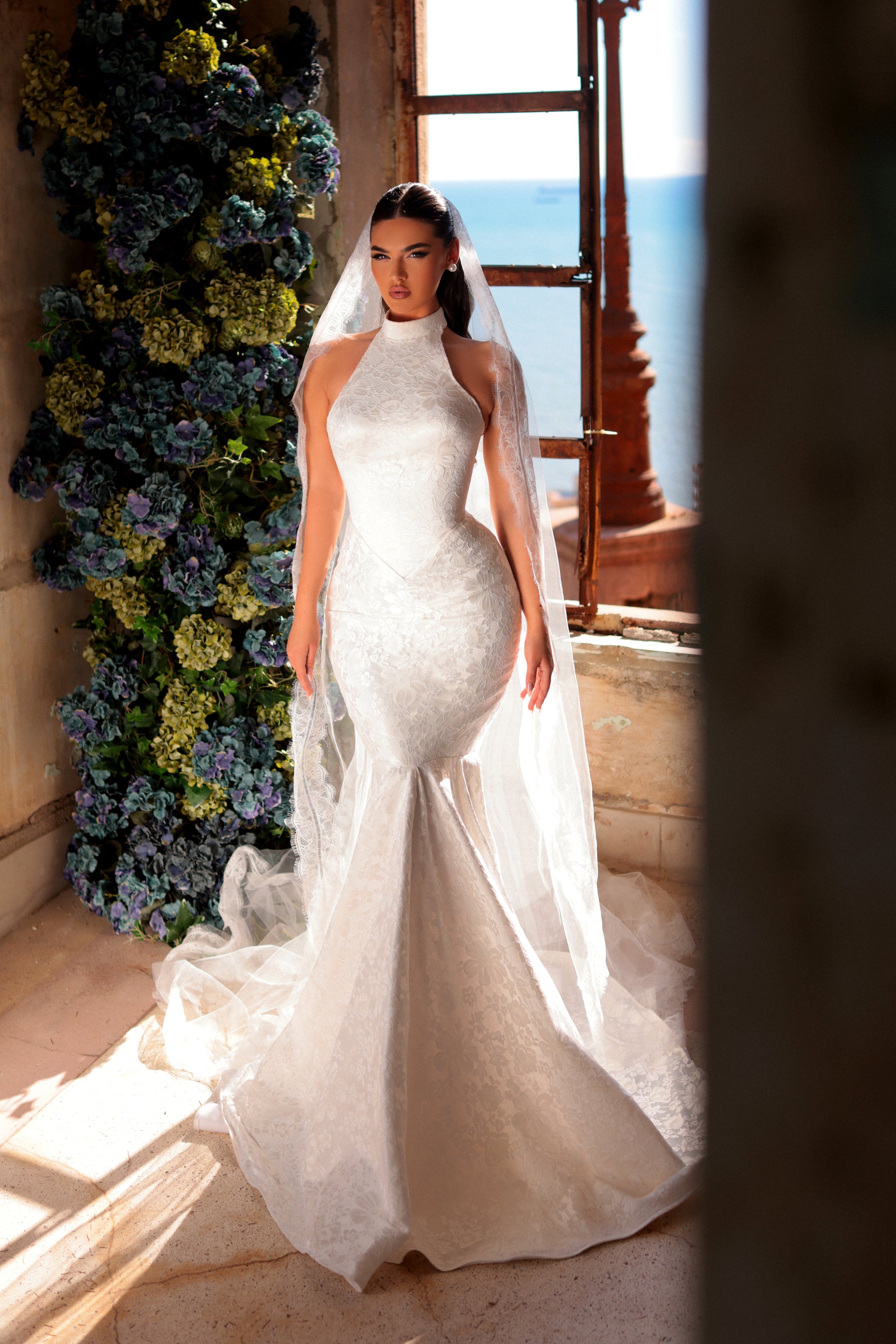 Woman in a white wedding dress standing in a room with floral decorations and a window view.